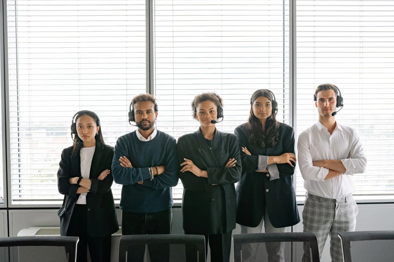 A diverse group of call center agents standing with arms crossed, wearing headsets in an office.